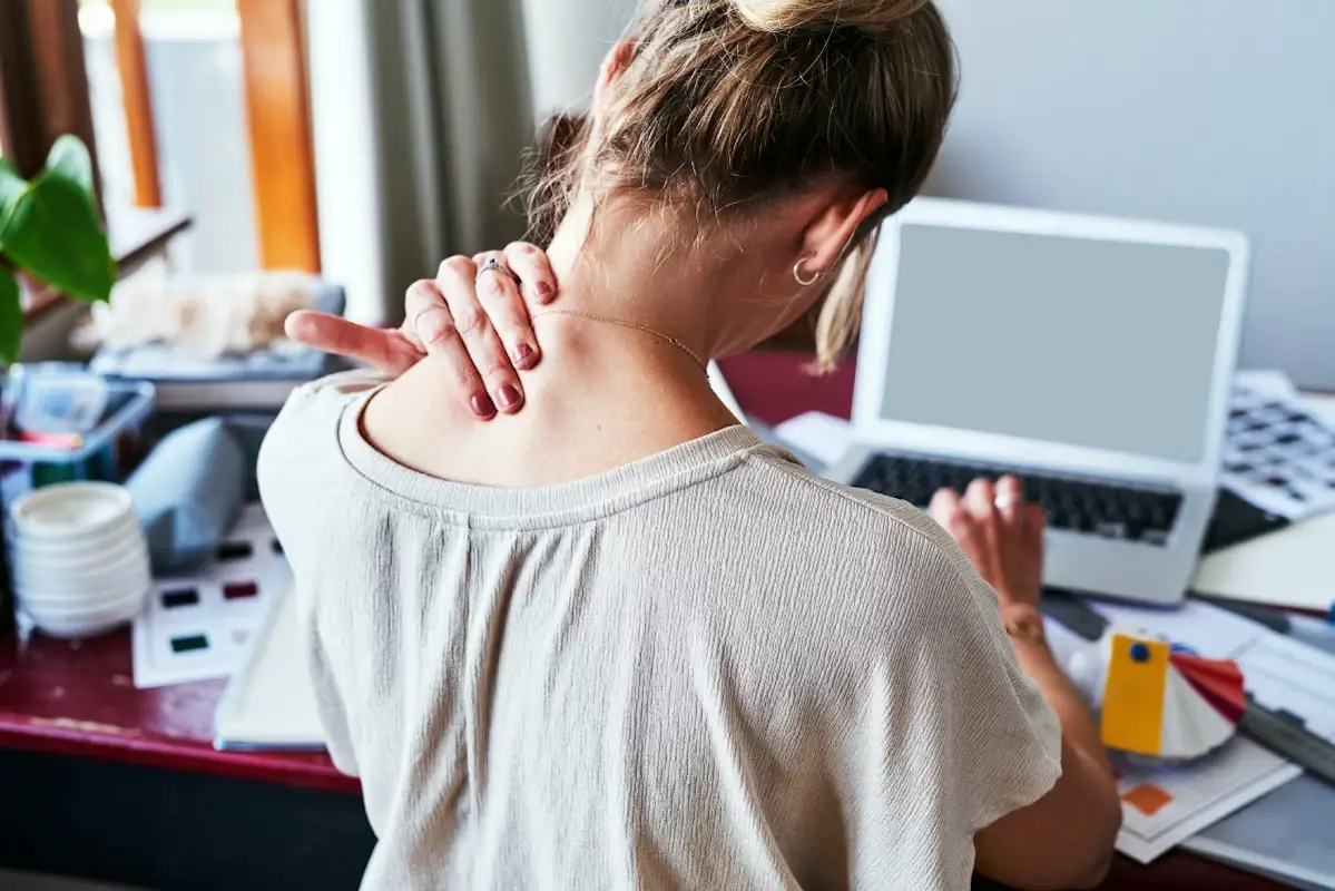 Mulher sentada, de costas, com a cabeça levemente inclinada, tocando a base do pescoço com a mão esquerda enquanto usa um laptop com a direita.